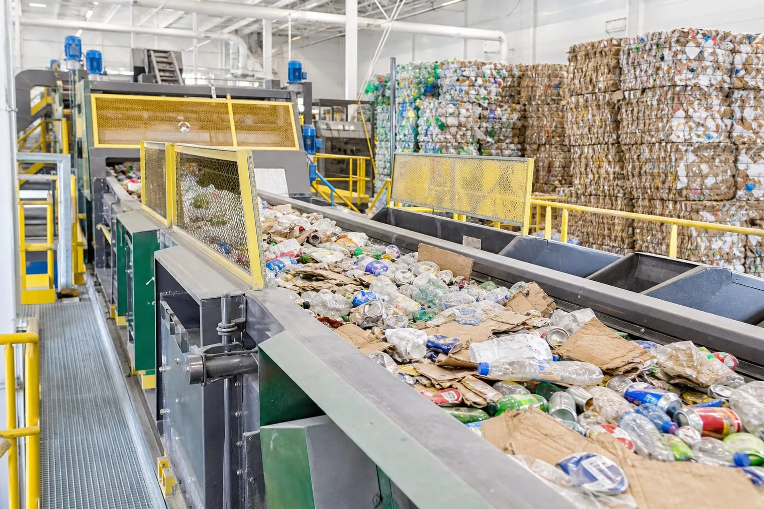 A materials recovery facility sorting line with cardboard, plastic bottles and cans moving along a conveyor belt past sorting screens, with baled commodities...