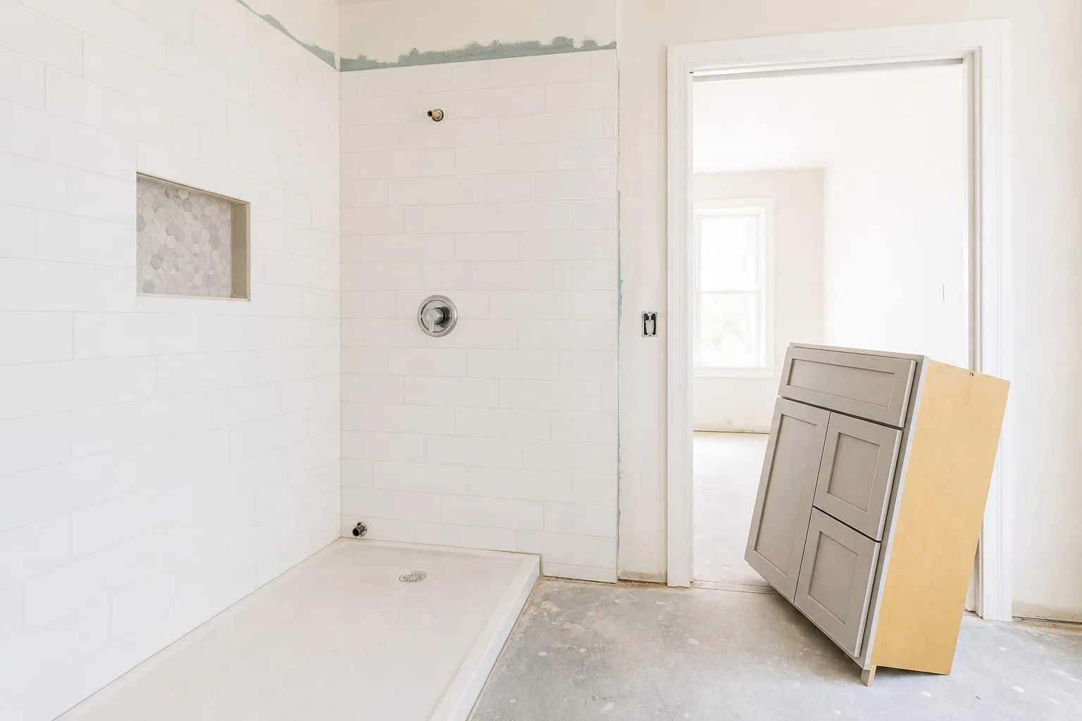 A half-finished bathroom remodel with tiled walls, an exposed shower valve, bare subfloor, and a boxed vanity leaning against the doorway, tools absent